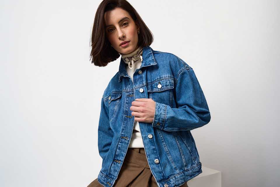 Mid-blue denim jacket styled over a white high-neck top with printed neck scarf, paired with brown tailored trousers for a smart casual everyday outfit.