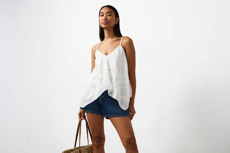 White embroidered camisole top with delicate lace detailing, styled with blue denim shorts, brown flat sandals and a woven straw tote bag for a casual summer holiday outfit.