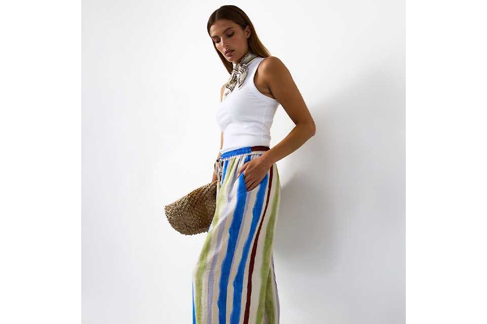 A woman wearing a white vest top, colourful striped wide leg trousers and black sandals.
