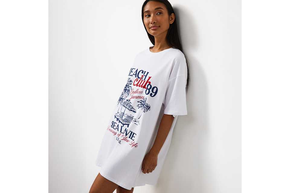 White oversized T-shirt nightdress featuring a Beach Club graphic print in blue and red with palm trees and seaside-inspired artwork.