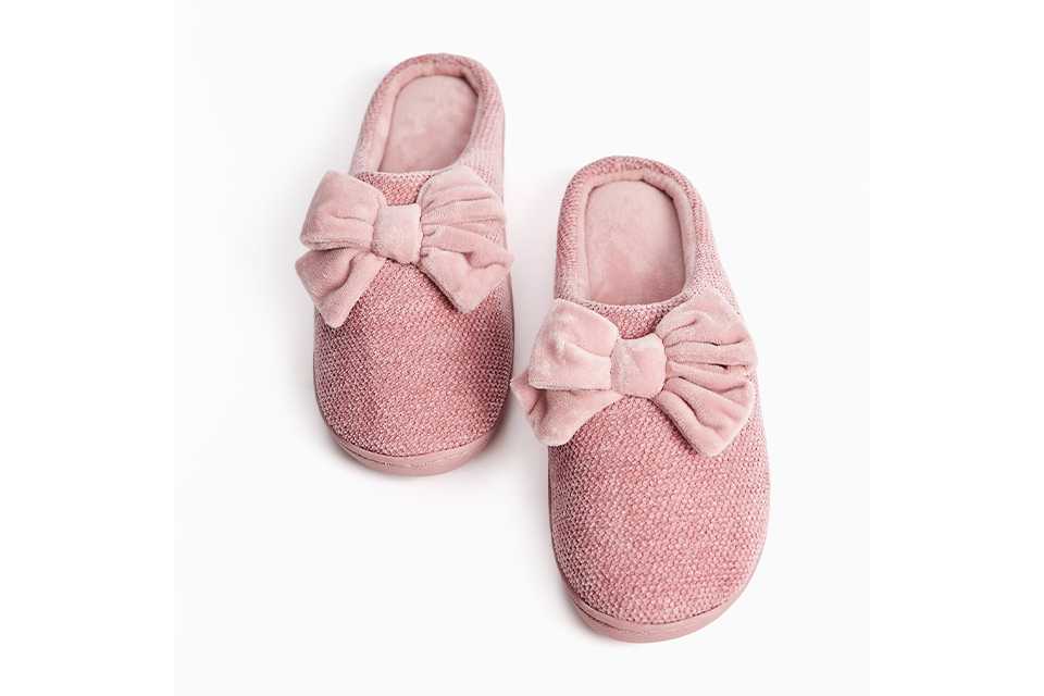 Pink textured mule slippers featuring oversized velvet bow detailing and a soft cushioned lining.