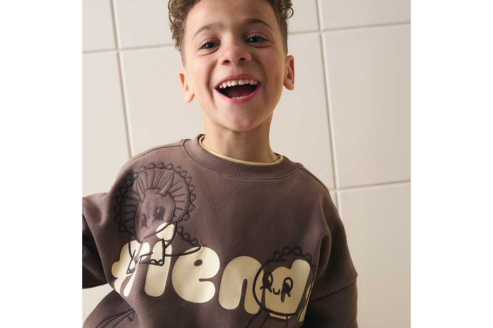 Close-up of a child wearing a brown graphic sweatshirt featuring playful dinosaur illustrations and bold cream lettering, photographed indoors against a neutral tiled background.