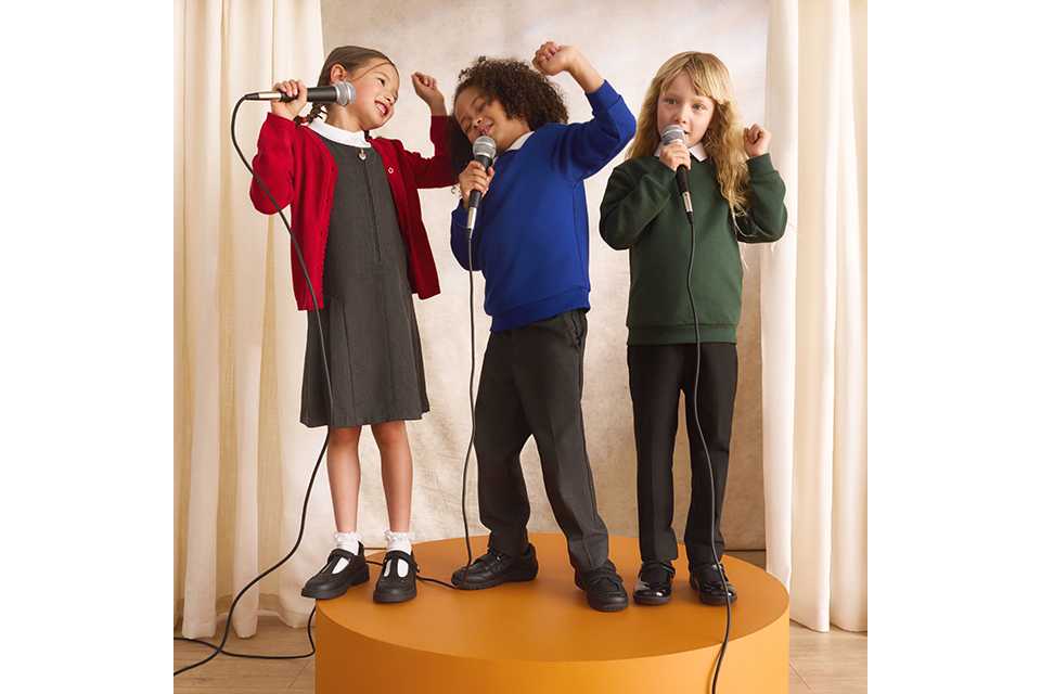 Three children standing on a stage platform singing into microphones, wearing colourful school uniform cardigans and jumpers during a school performance.