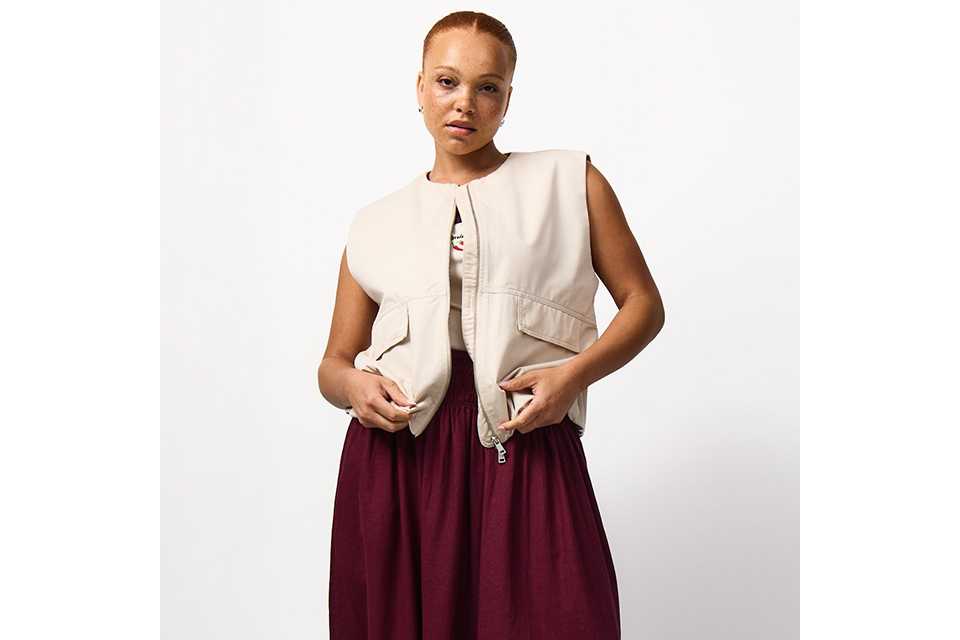 Burgundy maxi skirt styled with a cream sleeveless utility gilet featuring front flap pockets and a zip fastening for a contemporary casual look.