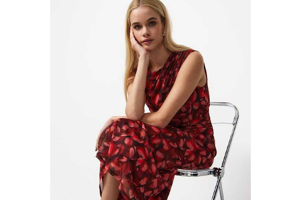 Red floral sleeveless midi dress with a sheer overlay, round neckline and flowing silhouette, styled with white sandals.