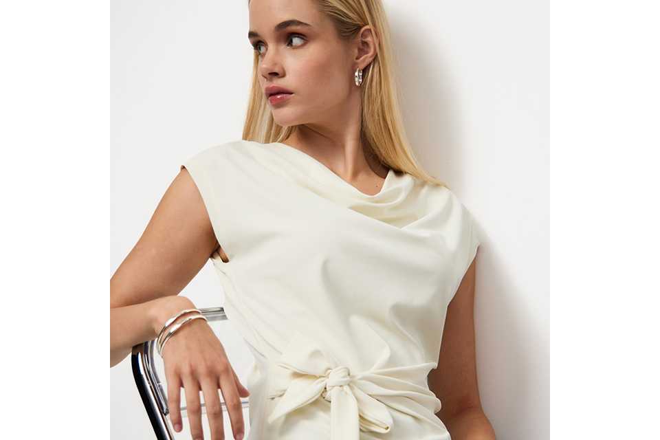 Ivory sleeveless blouse with a draped cowl neckline and side tie waist detail, styled with tailored trousers for an elegant contemporary look.