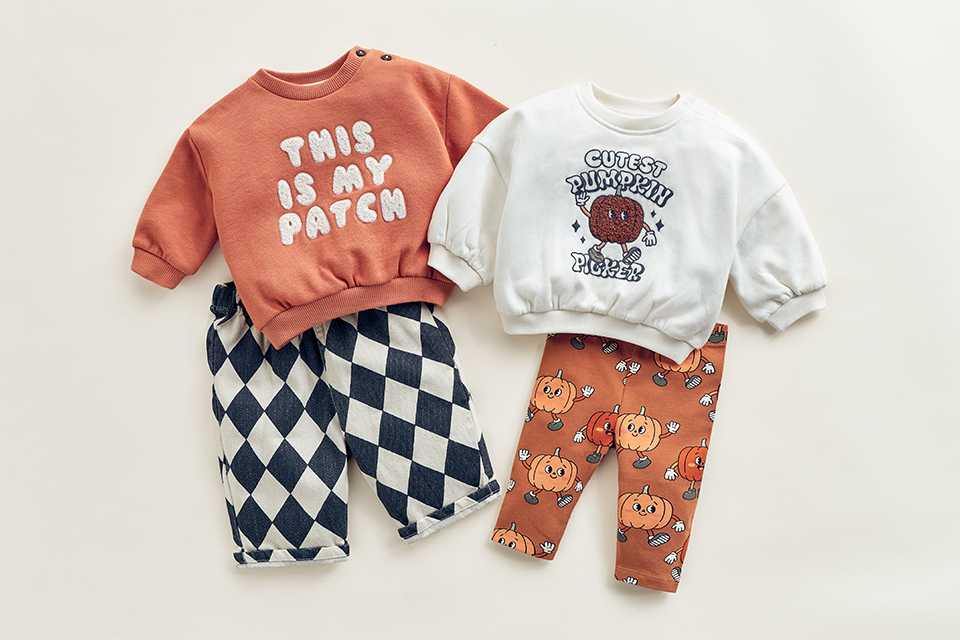 Baby Halloween outfit sets featuring pumpkin-themed sweatshirts paired with harlequin check trousers and pumpkin-print leggings, displayed in a flat-lay arrangement on a neutral background.