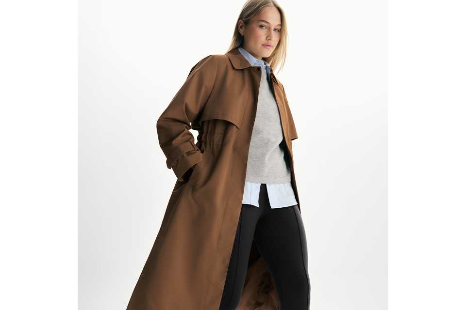 Brown trench coat outfit with grey sweater, blue shirt, black leggings, and black flats on a white background.