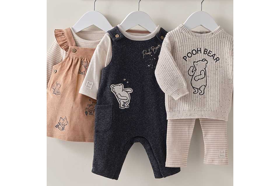 Disney Winnie the Pooh baby clothes collection featuring infant romper, knitted sweater, pinafore dress, and leggings in neutral beige, cream, and navy colours. Adorable newborn and baby outfit sets hanging on display.