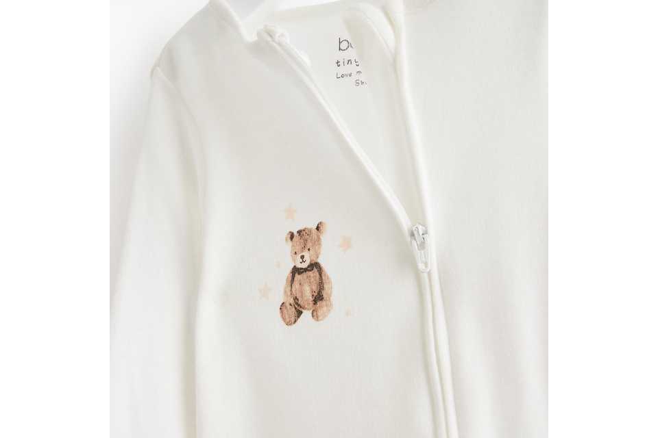 Cream baby zip-up sleepsuit featuring a teddy bear embroidery on the chest, made from soft cotton fabric and displayed on a hanger against a white background.