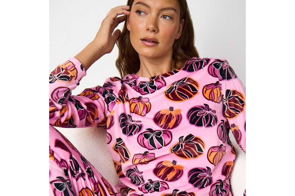 Person seated in a white textured armchair wearing a pink pajama set with an all-over pumpkin print, featuring a long-sleeve crew-neck top and matching lounge trousers against a light studio background.