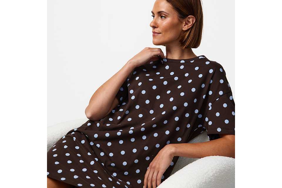 "Person seated in a white upholstered armchair wearing a brown short-sleeve midi dress with light blue polka dots, showcasing a relaxed contemporary fashion style against a neutral background.