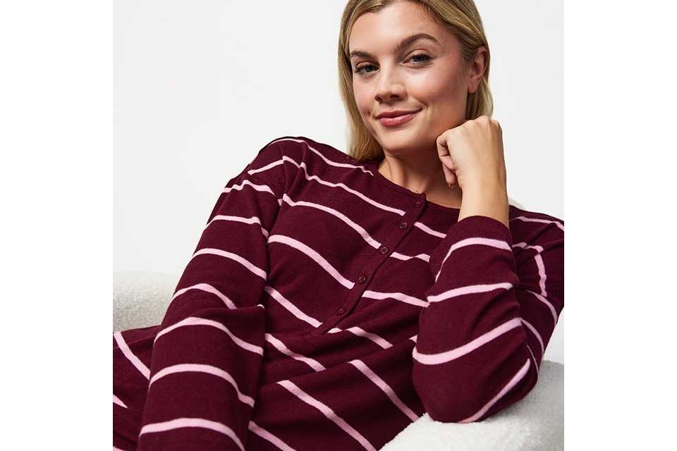 Person seated in a white textured armchair wearing a burgundy ribbed lounge set with a long-sleeve crew-neck top and matching wide-leg trousers against a neutral studio background.