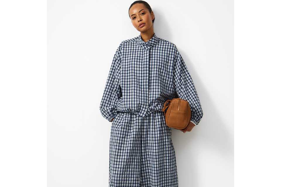 Blue and white check print shirt dress with long sleeves and gathered waist, styled with light blue sneakers and a tan crossbody bag in a minimalist studio setting.