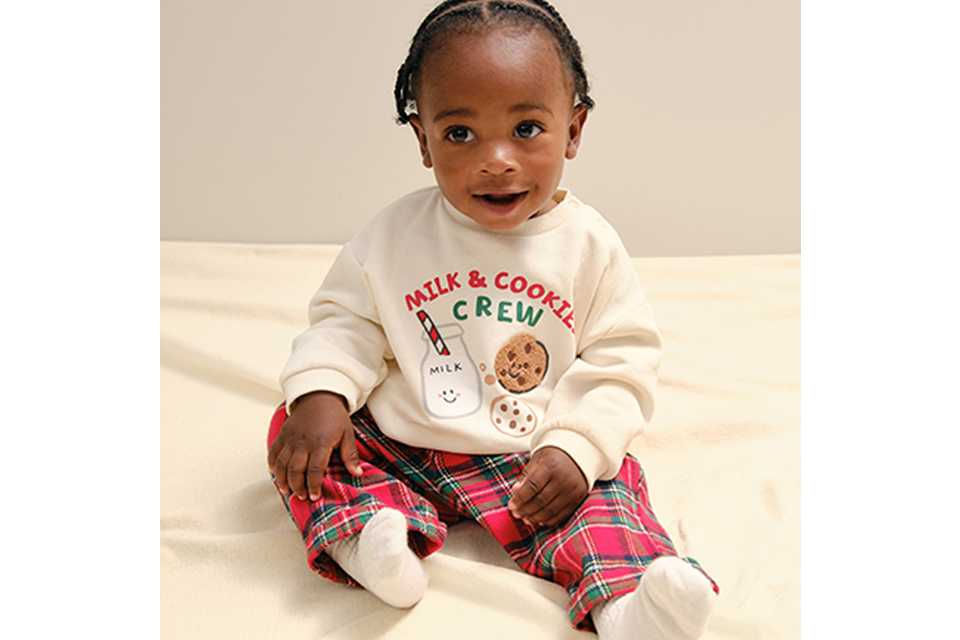 A baby wearing red tarten bottoms. with a cream jumper and a motif on it.