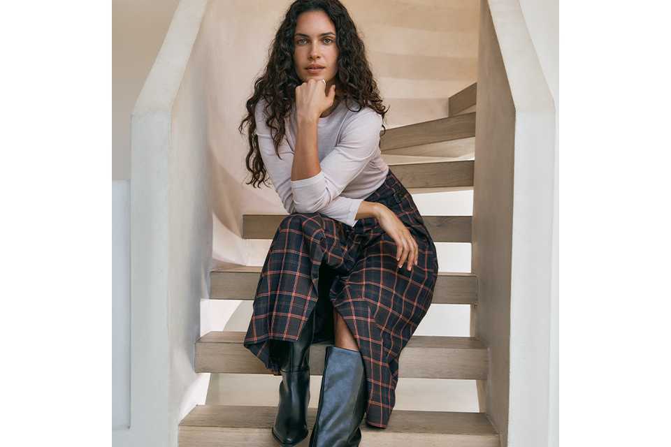 A woman wearing a neutral long sleeve top, with a black and red check skirt and black knee high boots.