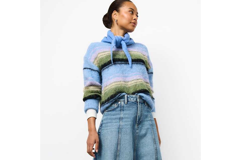 Light wash denim midi skirt styled with a soft blue striped knit sweater featuring a tie-neck detail, paired with cream ballet flats in a modern casual outfit.