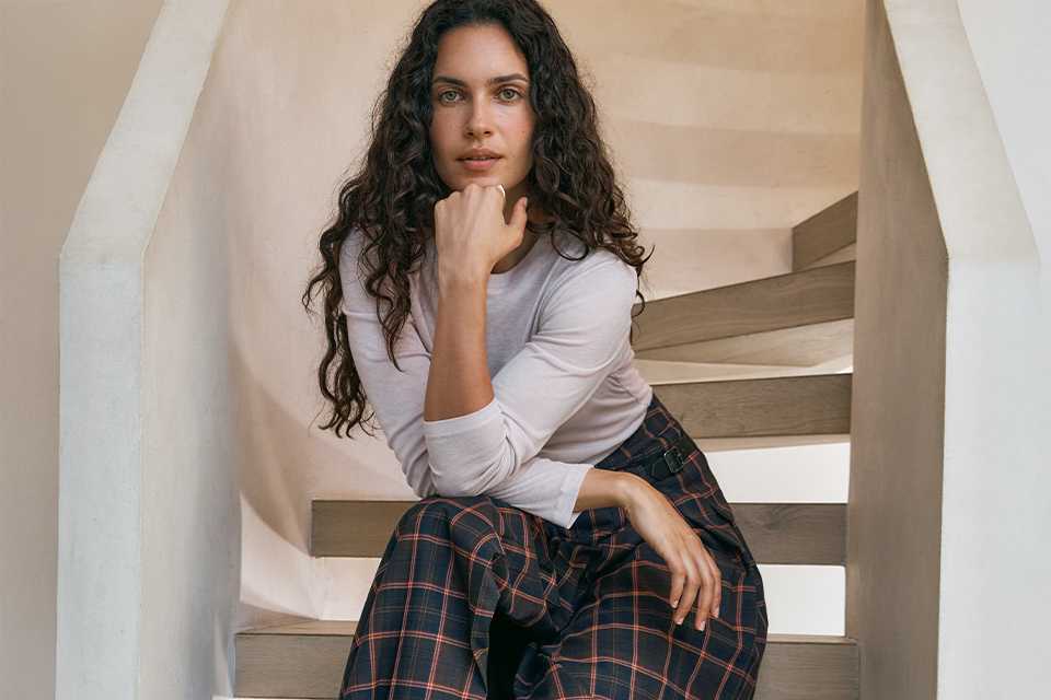 A woman wearing a neutral long sleeve top, with a black and red check skirt and black knee high boots