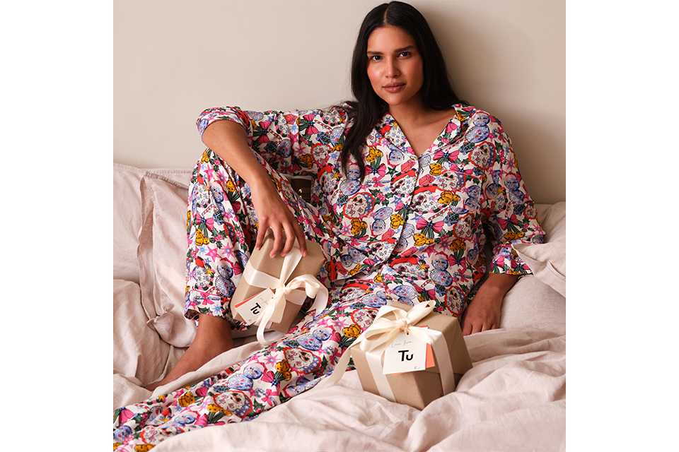 Model wearing colourful Christmas character print pyjamas sitting on a bed with wrapped gifts from Tu Clothing.