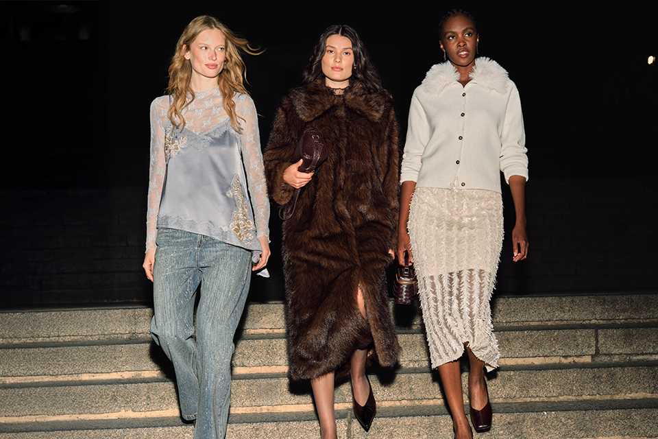 Three models showcasing luxury autumn-winter fashion on outdoor steps at night, featuring a lace-trim camisole with wide-leg trousers, a dark brown faux fur coat, and a textured knit cardigan paired with a fringed midi skirt.