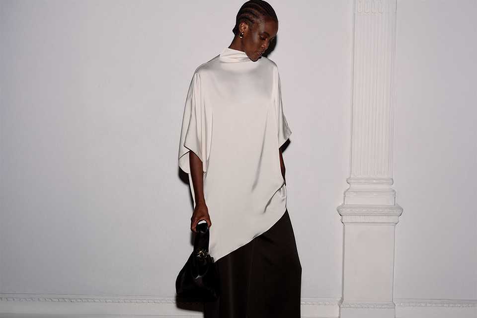 Women's cream draped high-neck top with asymmetric hem and cape-style sleeves, styled with wide-leg black trousers.