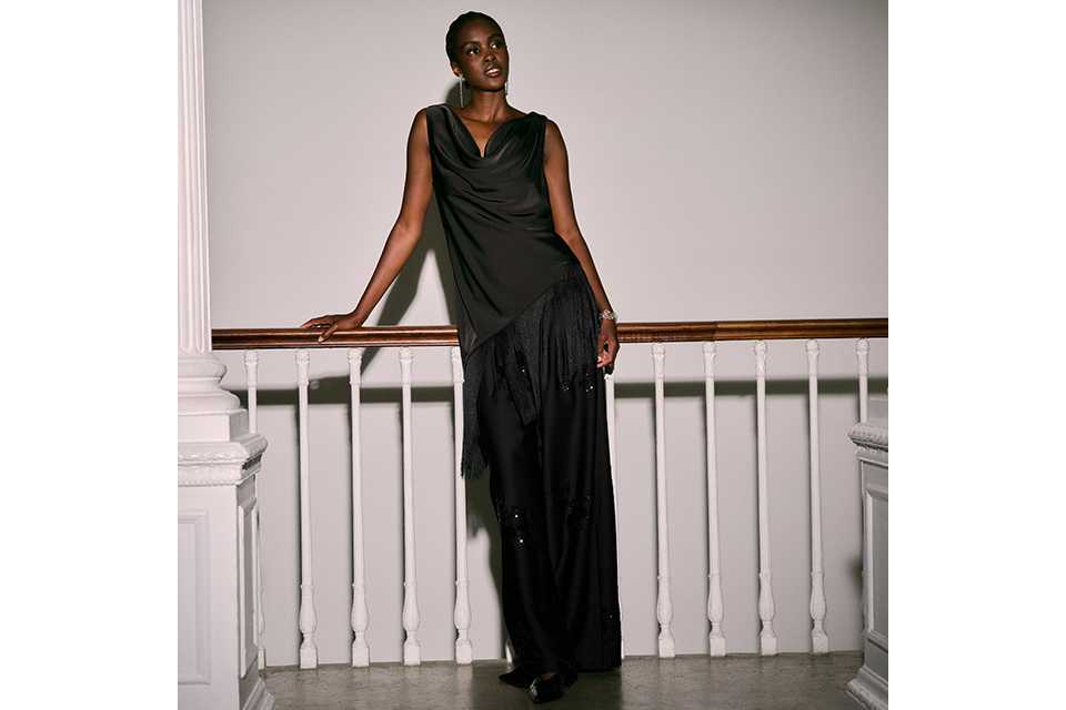 Women's black sleeveless draped top with tassel fringe hem and wide-leg black trousers, styled for eveningwear.