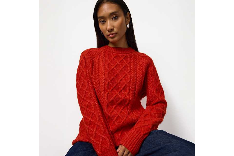 Bright red cable-knit crew neck sweater featuring an intricate textured pattern, styled with dark blue denim jeans for a classic casual winter fashion look.