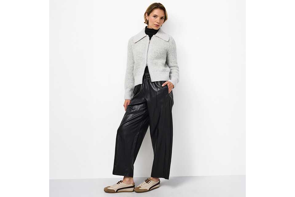 Light grey zip-front knit cardigan with a wide collar, layered over a black turtleneck and paired with black wide-leg faux leather trousers and casual low-top sneakers for a contemporary smart-casual look.