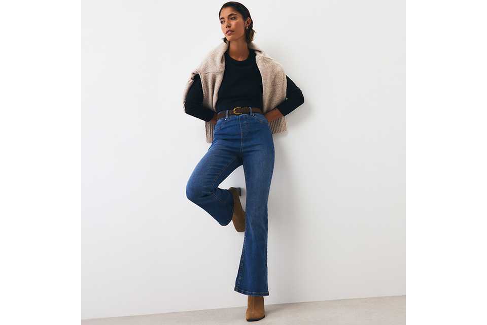 Blue high-waisted bootcut jeans paired with a black long-sleeve top, brown belt, ankle boots, and a beige knit sweater draped over the shoulders for a casual smart-casual look.