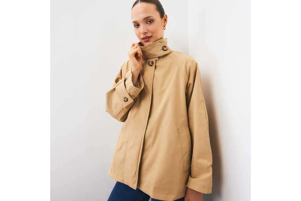 Beige high-collar utility jacket featuring oversized sleeves, button-detail cuffs, concealed front fastening, and patch pockets, styled with blue denim jeans in a minimalist studio setting.