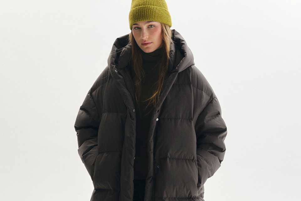 Long dark brown quilted puffer coat with an oversized hood, styled with a lime green knit beanie, black outfit, and chunky ankle boots for cold-weather winter fashion.