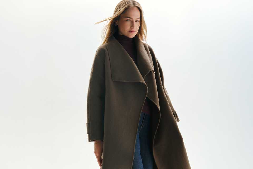 Long dark brown wrap coat with an oversized draped collar, layered over a dark turtleneck top and blue denim jeans for an elegant autumn and winter outerwear look.