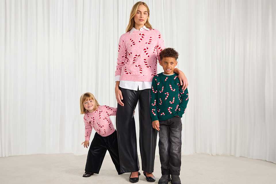 Three people wearing festive sweaters with candy cane patterns, standing in front of a white curtain backdrop. Two sweaters are pink with red candy canes, and one sweater is green with red candy canes.