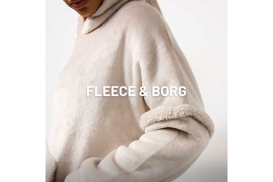 Close-up of a person wearing a light beige, plush fleece hoodie with a soft, textured finish. The garment features a cosy hood and a thick, sherpa-like trim detail on the sleeve. The person’s hand is partially tucked into a front pocket.