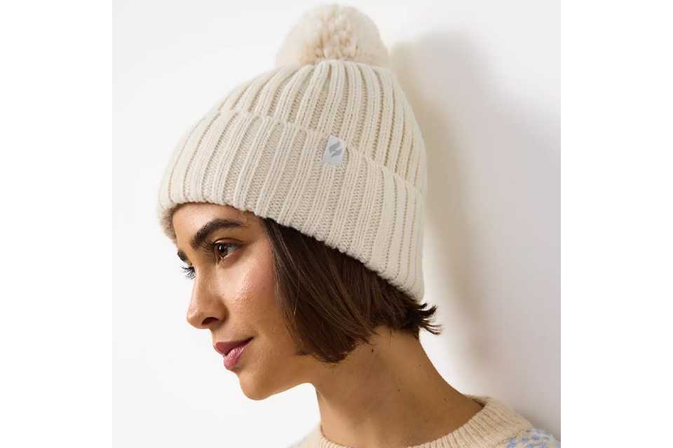 Close-up of a person wearing a cream-colored knitted winter hat with a pom-pom on top, paired with a light beige sweater featuring a textured pattern.