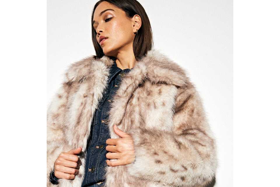 Person dressed in a thick, light brown and cream faux fur coat layered over a dark textured top, shown from the waist up.