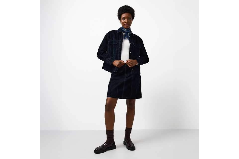 Person wearing a dark denim jacket and matching denim skirt, styled with a white top and black ankle boots, standing against a minimal white backdrop.