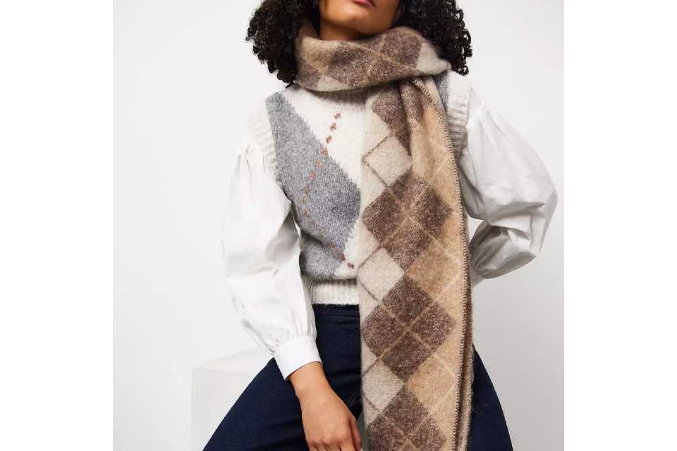 Person wearing a thick, oversized checkered scarf in shades of beige, gray, and white, draped over a white long-sleeve top.
