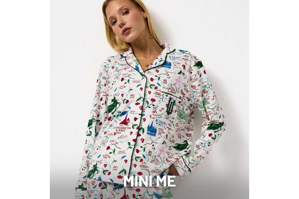 Person wearing a white pajama set with colorful illustrated prints, including castles, trees, and text. The top is button-up with dark piping along the edges, and the pants have a matching pattern. The background is plain white.