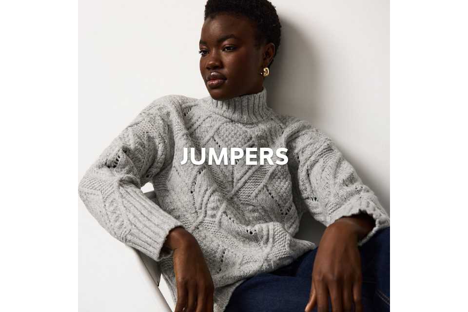 Person seated on a white chair wearing a light gray cable-knit sweater with a high neckline and dark blue jeans.