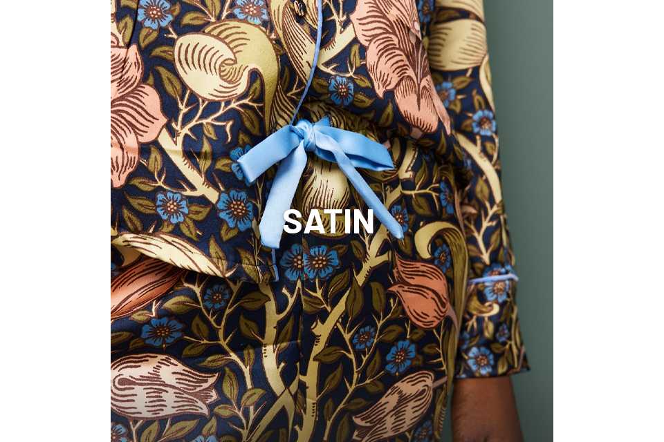 Close-up of a person wearing a dark satin pyjama set with an intricate floral and bird print in shades of gold, peach, and blue, featuring a light blue ribbon tied at the waist.