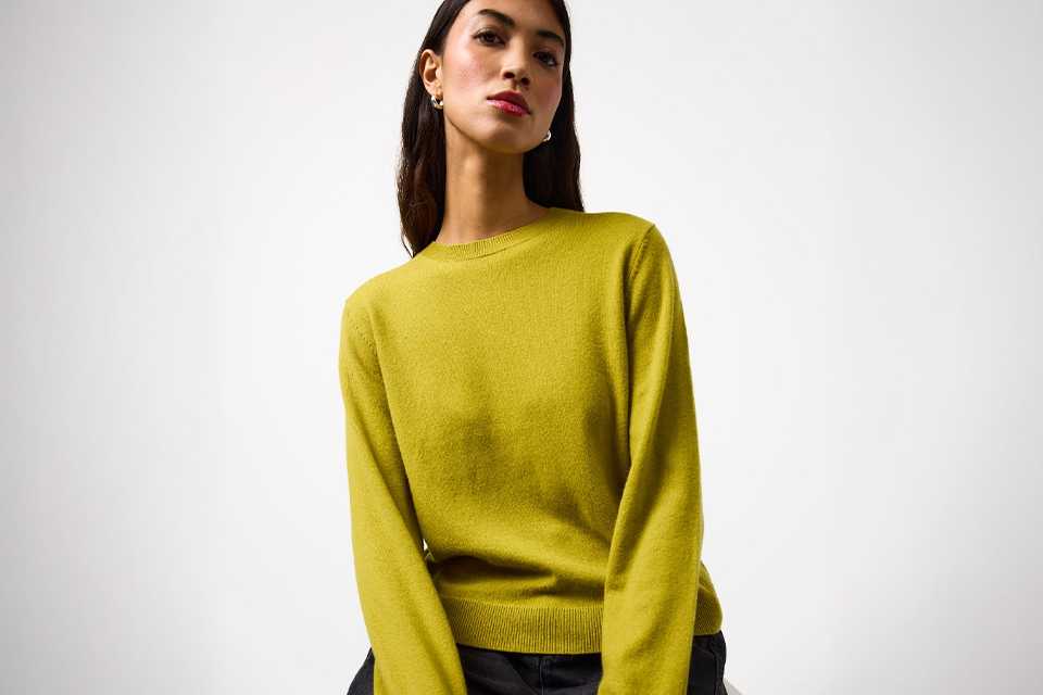 Person wearing a bright yellow long-sleeve crew-neck sweater paired with dark grey jeans, seated on a light-colored surface.