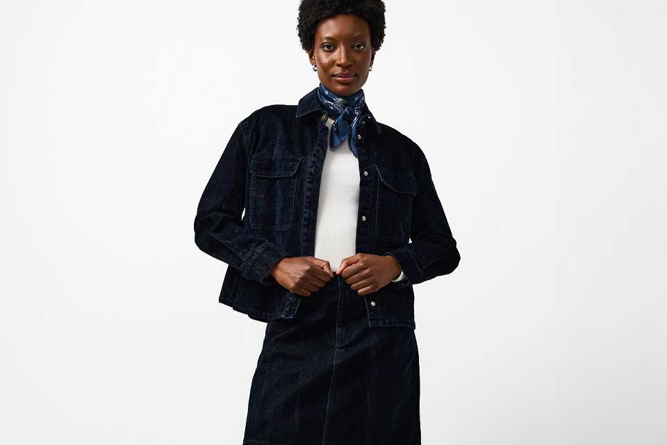 Person wearing a dark denim jacket and matching mini skirt, styled with a white top, blue patterned neck scarf, brown socks, and black loafers.