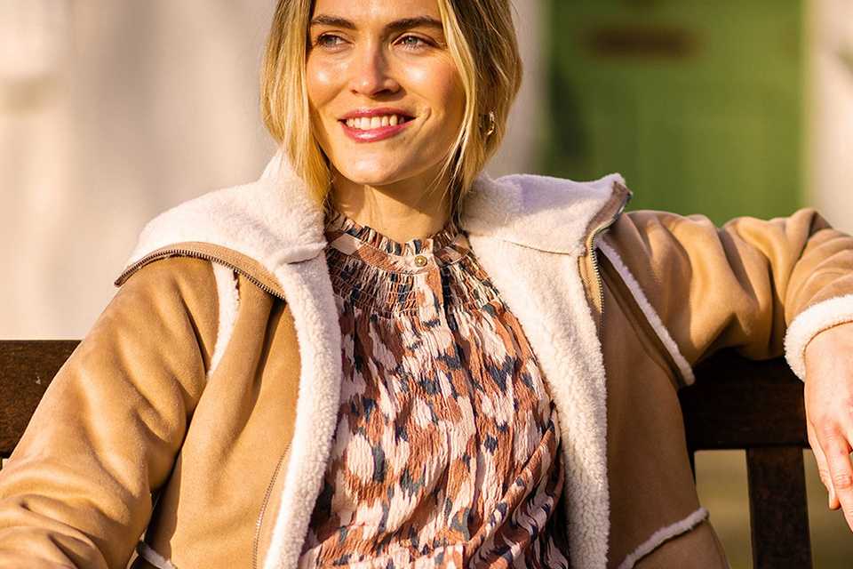 Person sitting on a wooden bench wearing a tan shearling-lined jacket over a patterned blouse, paired with high-waisted light blue jeans and holding a cream textured bag.