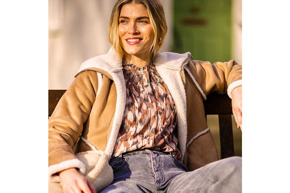 Person sitting on a wooden bench wearing a tan shearling-lined jacket over a patterned blouse, paired with high-waisted light blue jeans and holding a cream textured bag.