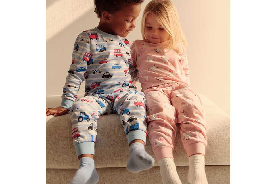 Two children sitting on a light-colored sofa wearing cozy cotton pajamas; one in a blue set with colorful vehicle prints and the other in a pink set with delicate animal patterns, perfect for bedtime comfort.
