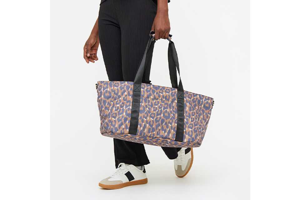 Person wearing a black textured top and matching pants, holding a large rectangular baby changing bag with a blue and brown leopard print design and black shoulder straps. The person is also wearing white sneakers with black and tan accents.