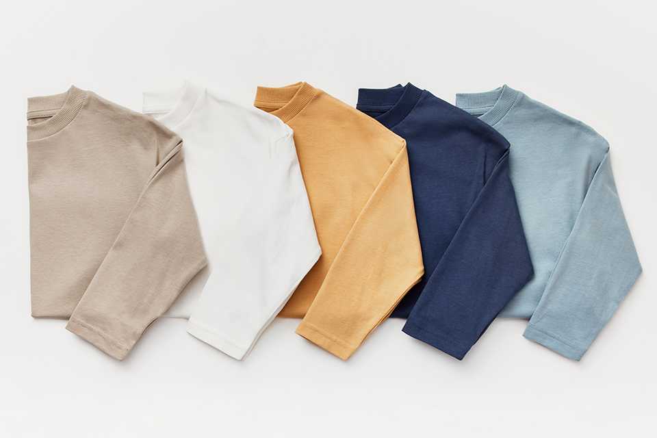 Five long-sleeve tops in neutral and muted tones—beige, white, mustard, navy, and light blue—arranged in a row.