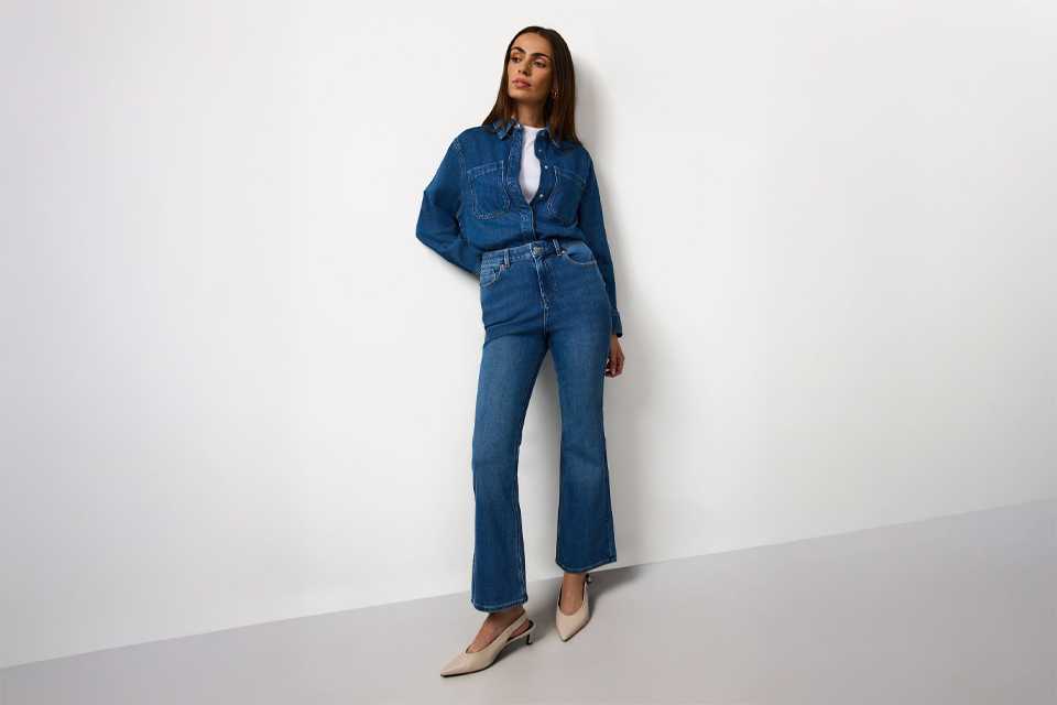 Person wearing a dark denim jacket and matching mini skirt, styled with a white top, blue patterned neck scarf, brown socks, and black loafers.