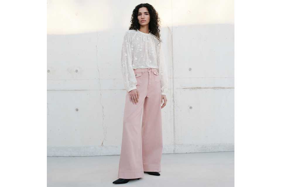 Person standing against a light concrete wall wearing a long-sleeved white lace blouse paired with high-waisted, wide-leg pink trousers and black pointed shoes.